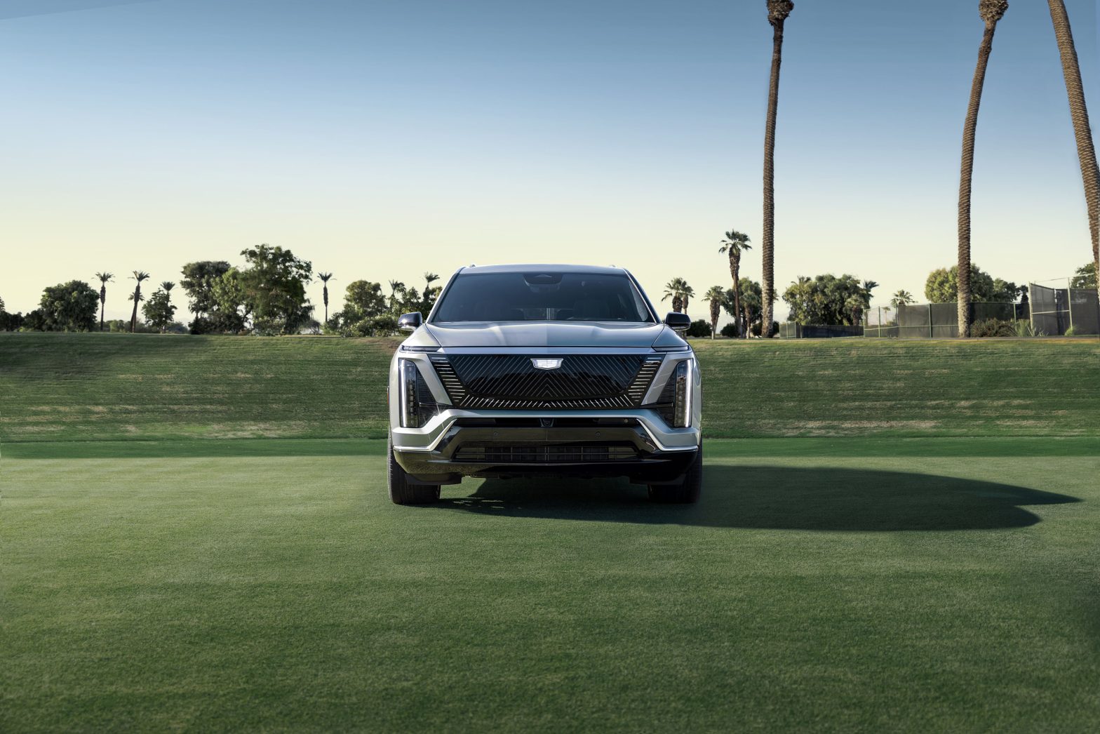 Front view of the 2026 Cadillac Vistiq electric SUV with Black Crystal Shield grille, displayed on a green field with palm trees in the background.