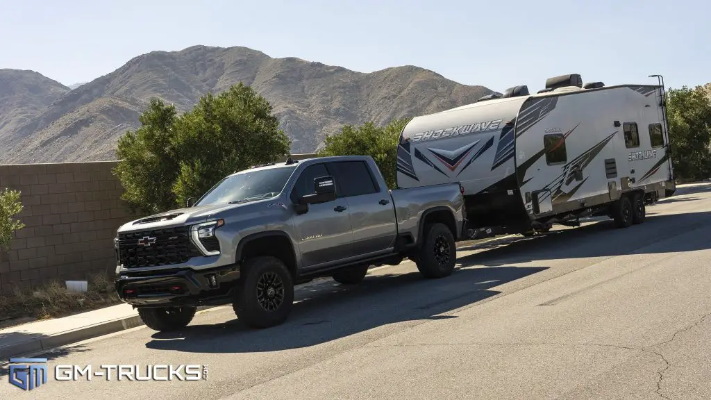 Silver Chevrolet Silverado 2500 HD towing a large Shockwave travel trailer on a desert road with mountains in the background.