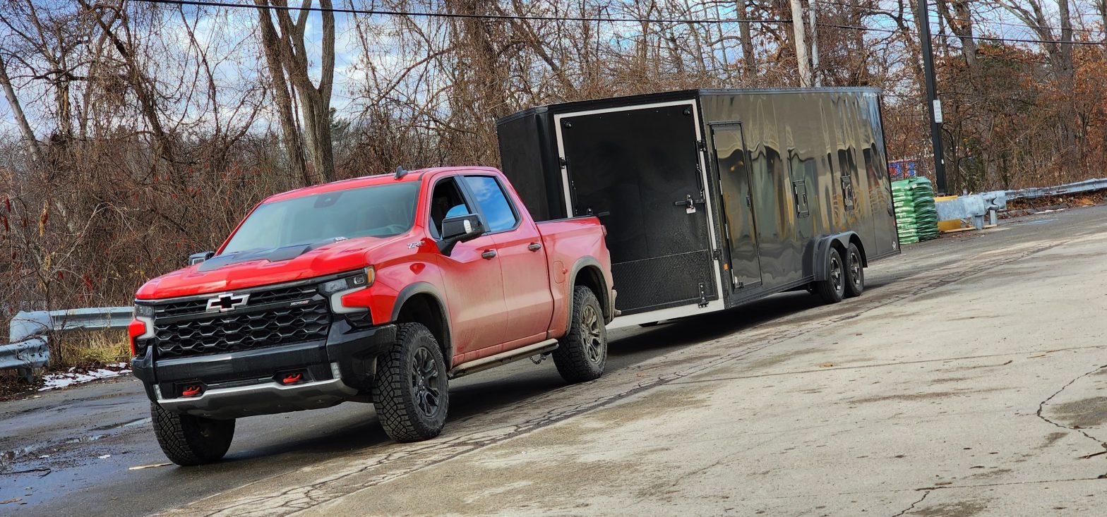 Red Chevrolet Silverado 1500 ZR2 towing a large black enclosed trailer on a wet road